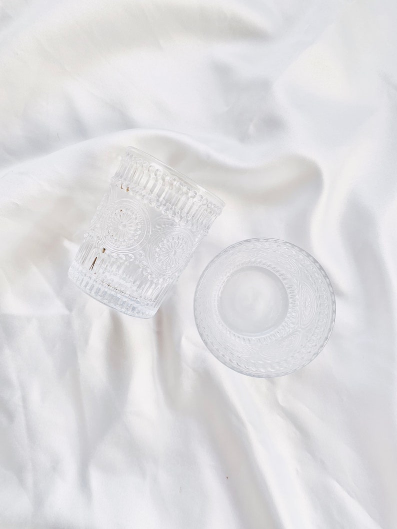 May include: Two clear glass drinking glasses with embossed floral patterns. One glass is upright, and the other is on its side. The glasses are on a white, textured surface. The glasses are likely for water or other beverages.