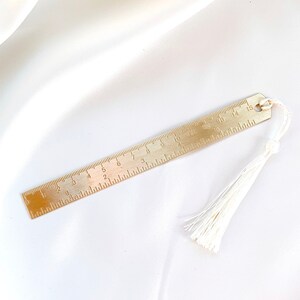 May include: A brass bookmark with a ruler design, featuring inch measurements. The bookmark has a small loop at the top and a white tassel attached. The ruler is marked with numbers from 1 to 15.