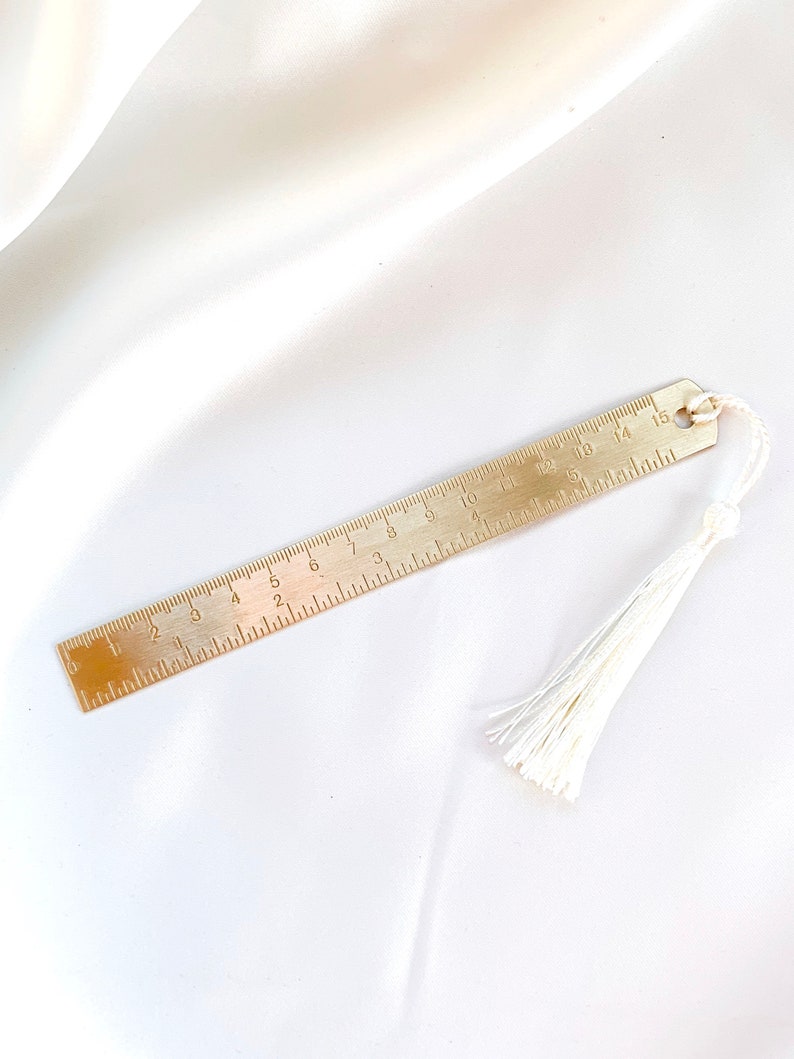 May include: A brass bookmark with a ruler design, featuring inch measurements. The bookmark has a small loop at the top and a white tassel attached. The ruler is marked with numbers from 1 to 15.
