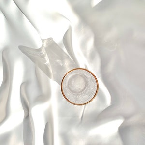 May include: A clear glass with a gold rim sits on a white, silky fabric. The glass has an etched design and is empty. The lighting creates shadows and highlights on the fabric, adding depth to the image.