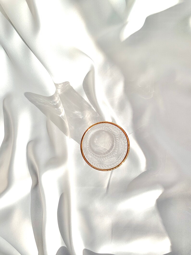 May include: A clear glass with a gold rim sits on a white, silky fabric. The glass has an etched design and is empty. The lighting creates shadows and highlights on the fabric, adding depth to the image.