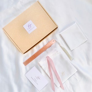 May include: An overhead shot of a collection of items on a white satin surface. Includes a tan box with a logo, a rose gold ruler, a white pouch with a pink ribbon, a small white card, and a white pouch with a tassel.