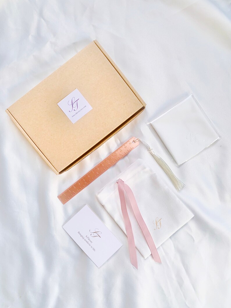 May include: An overhead shot of a collection of items on a white satin surface. Includes a tan box with a logo, a rose gold ruler, a white pouch with a pink ribbon, a small white card, and a white pouch with a tassel.