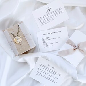 May include: A wrapped gift with a wax seal and several white cards with text on a white satin background. The cards describe a handcrafted water glass with a golden ring and care instructions. The gift is tied with twine and dried sprigs.
