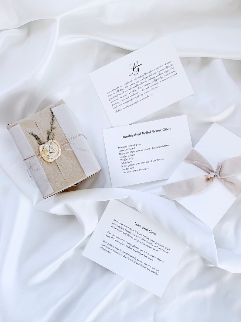 May include: A wrapped gift with a wax seal and several white cards with text on a white satin background. The cards describe a handcrafted water glass with a golden ring and care instructions. The gift is tied with twine and dried sprigs.