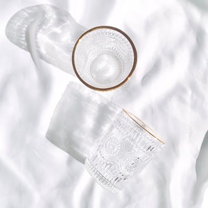 May include: Two clear drinking glasses with intricate embossed patterns and gold rims are displayed on a white, textured surface. The glasses cast shadows, enhancing the play of light and shadow. The design of the glasses is vintage-inspired.