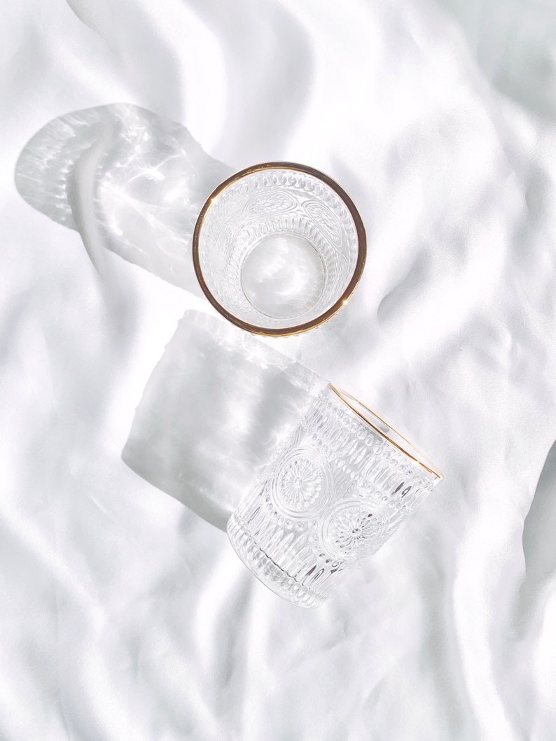 May include: Two clear drinking glasses with intricate embossed patterns and gold rims are displayed on a white, textured surface. The glasses cast shadows, enhancing the play of light and shadow. The design of the glasses is vintage-inspired.