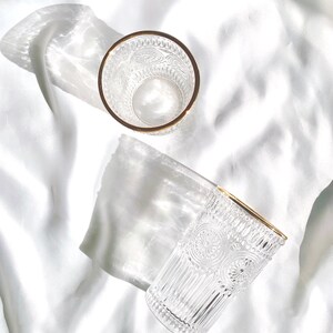 May include: Two clear drinking glasses with intricate embossed patterns and gold rims are displayed on a white, textured surface. The glasses are positioned at different angles, showcasing their design and the play of light and shadow.