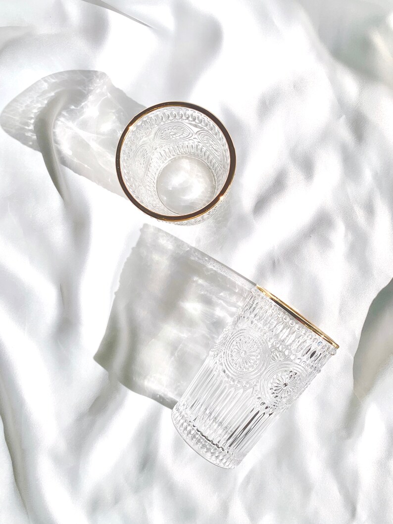May include: Two clear drinking glasses with intricate embossed patterns and gold rims are displayed on a white, textured surface. The glasses are positioned at different angles, showcasing their design and the play of light and shadow.