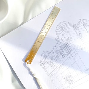 May include: A brass ruler with inch markings and a white tassel bookmark rests on a white paper with a pencil sketch of a building. The ruler is 6 inches long. The background is a white, silky fabric.