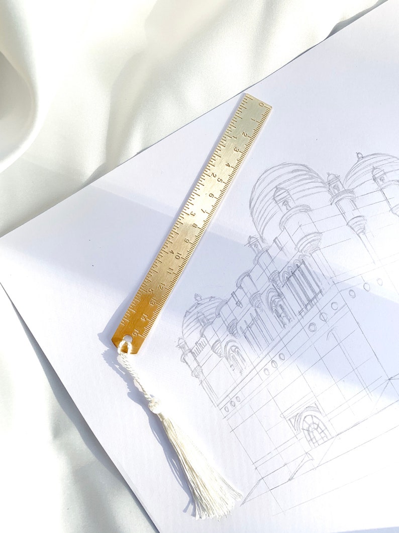 May include: A brass ruler with inch markings and a white tassel bookmark rests on a white paper with a pencil sketch of a building. The ruler is 6 inches long. The background is a white, silky fabric.