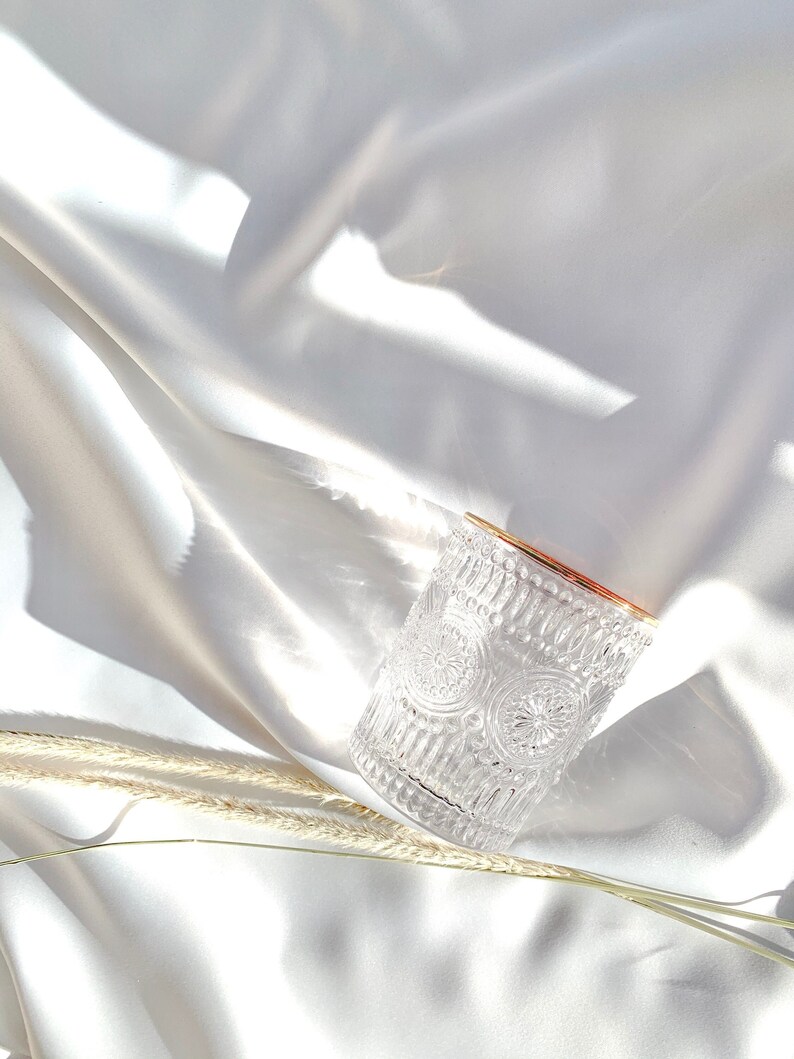 May include: A clear, patterned glass with a gold rim sits on a white, silky surface. The glass features an intricate design of repeating circular patterns. Dried wheat stalks are placed near the glass, adding a natural element to the composition.
