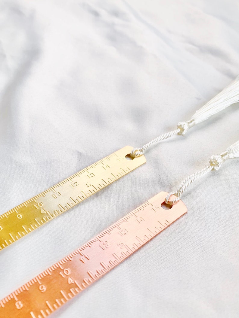 May include: Two metal bookmarks with ruler markings and white tassels. One bookmark is gold-toned, the other is rose gold. Each bookmark has measurements from 8 to 15 inches.