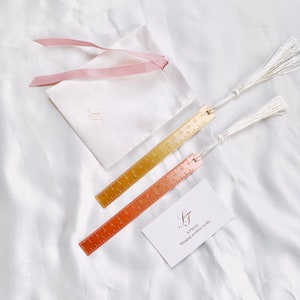May include: Two metal ruler bookmarks with tassels, one gold and one copper, next to a white pouch with a pink ribbon and a card that reads "S.Theory Bringing aesthetic to life."
