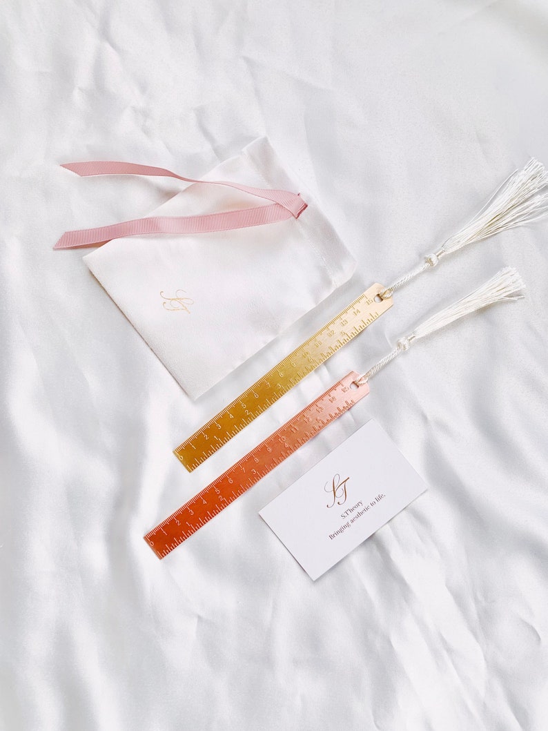 May include: Two metal ruler bookmarks with tassels, one gold and one copper, next to a white pouch with a pink ribbon and a card that reads "S.Theory Bringing aesthetic to life."