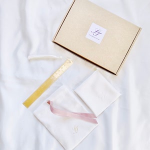 May include: A gift set featuring a tan cardboard box with a logo, a gold metal bookmark with ruler markings, two white pouches with pink ribbon and gold initials, and a white handkerchief with gold initials. A white tassel is attached to the bookmark.
