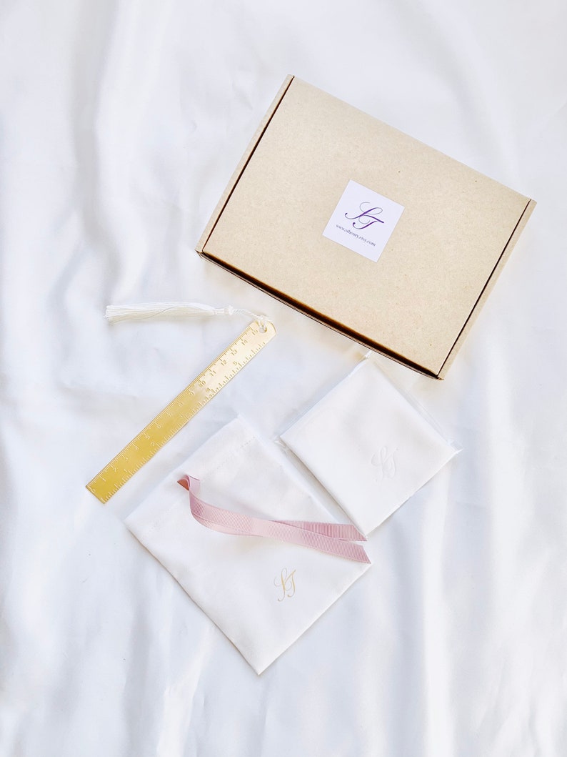 May include: A gift set featuring a tan cardboard box with a logo, a gold metal bookmark with ruler markings, two white pouches with pink ribbon and gold initials, and a white handkerchief with gold initials. A white tassel is attached to the bookmark.