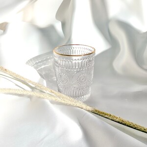 May include: Clear glass tumbler with an intricate embossed pattern and a gold rim. The glass is set against a backdrop of white fabric, with dried wheat stalks in the foreground. The design features repeating floral and geometric shapes.