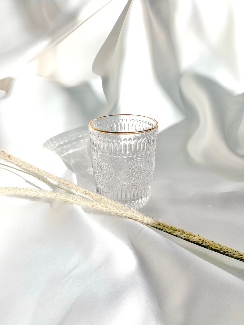 May include: Clear glass tumbler with an intricate embossed pattern and a gold rim. The glass is set against a backdrop of white fabric, with dried wheat stalks in the foreground. The design features repeating floral and geometric shapes.