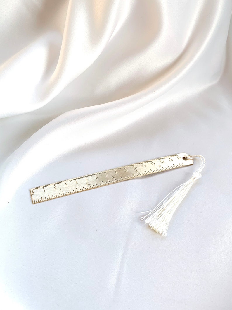 May include: A gold-colored metal bookmark with ruler markings and a white tassel. The bookmark is lying on a white satin fabric. The ruler markings are in inches.
