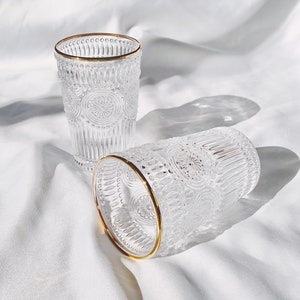 May include: Two clear glass tumblers with a gold rim. The glasses have a textured, embossed design with a floral pattern. One glass is upright, and the other is lying on its side. The background is a white, textured fabric.