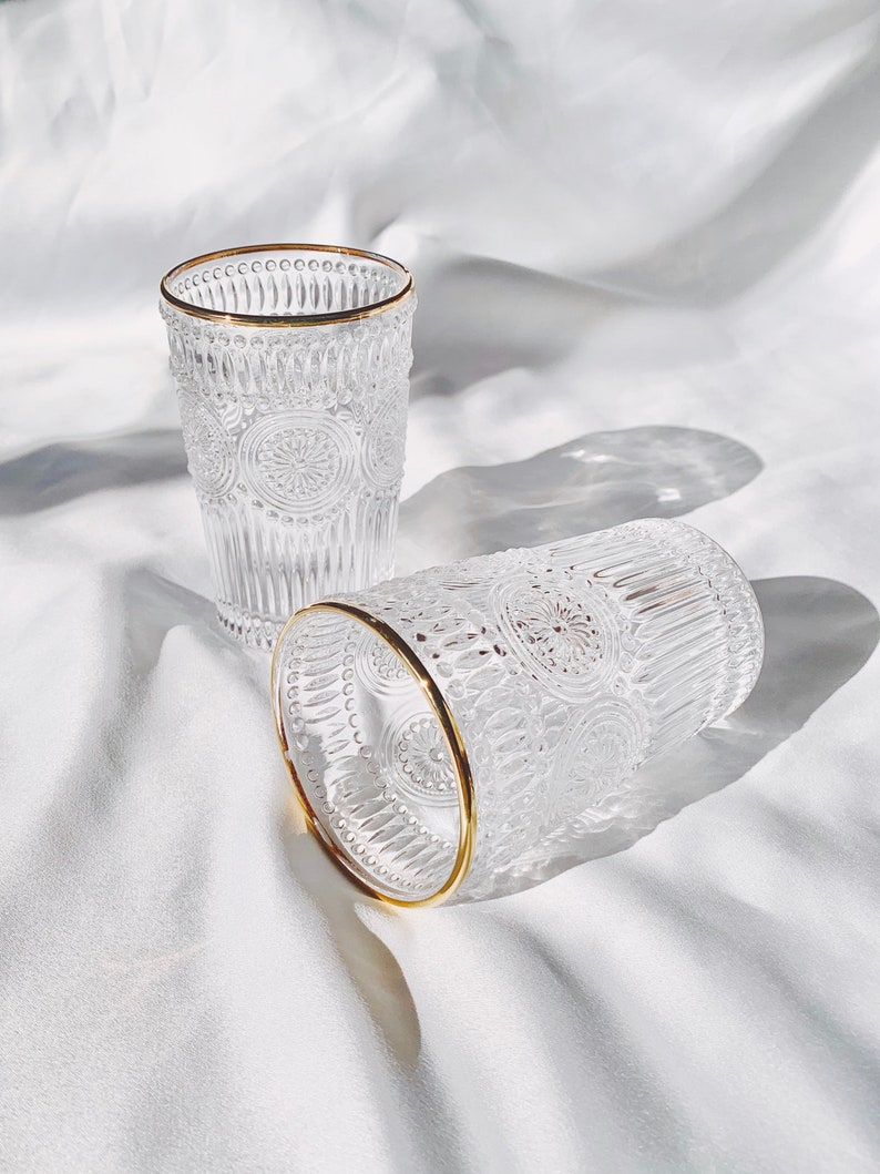 May include: Two clear glass tumblers with a gold rim. The glasses have a textured, embossed design with a floral pattern. One glass is upright, and the other is lying on its side. The background is a white, textured fabric.