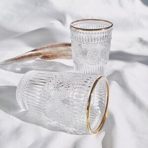 May include: Two clear drinking glasses with intricate embossed patterns and gold rims. The glasses are cylindrical and sit on a white surface, with one glass lying on its side. The design features floral and geometric elements.