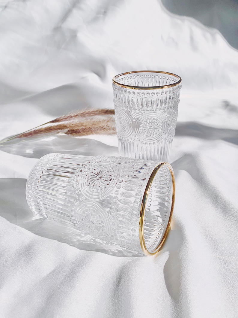 May include: Two clear drinking glasses with intricate embossed patterns and gold rims. The glasses are cylindrical and sit on a white surface, with one glass lying on its side. The design features floral and geometric elements.