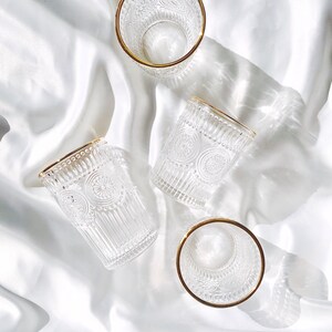 May include: Four clear drinking glasses with intricate embossed designs and gold rims are arranged on a white, silky surface. The glasses are empty, and the light creates shadows, highlighting the details of the glassware.