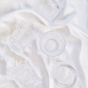 May include: Clear glass tumblers with embossed floral and vertical line patterns. The glasses are arranged on a white, silky fabric, creating a delicate and elegant display. The design of the glasses adds a vintage touch.