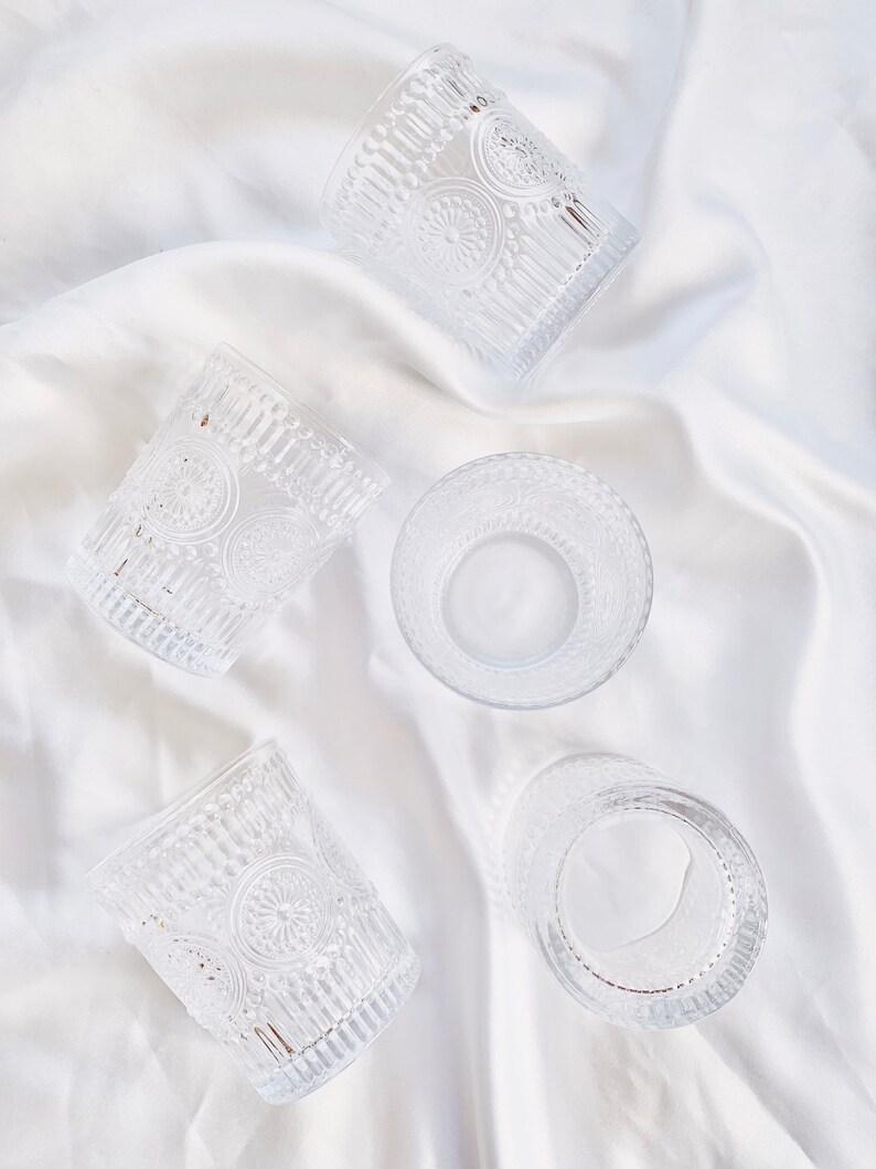 May include: Clear glass tumblers with embossed floral and vertical line patterns. The glasses are arranged on a white, silky fabric, creating a delicate and elegant display. The design of the glasses adds a vintage touch.