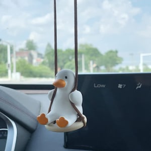 Peut inclure: Une figurine de canard blanc avec un bec et des pieds orange, suspendue à une ficelle brune, est sur une balançoire. Le canard est assis sur un siège en bois. La balançoire est suspendue au rétroviseur d'une voiture.