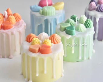 May include: Assortment of colorful mini cake candles with various toppings, including hearts, flowers, and swirls. The candles are drizzled with white icing and have a pastel color scheme.