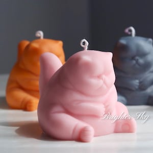 May include: Five hand-poured candles shaped like cats in various colours, including white, orange, pink, grey, and purple. The candles are sitting on a white surface.