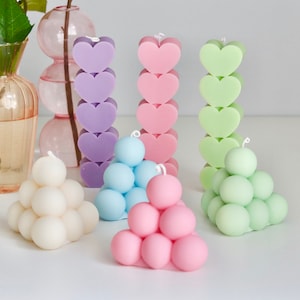 Puede incluir: Surtido de velas coloridas en varias formas, incluyendo una pila de corazones y un grupo de esferas. Las velas están dispuestas sobre una superficie blanca.
