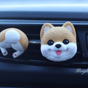 May include: Two car air freshener ornaments shaped like a Shiba Inu dog. One ornament is a front-facing view of the dog's face, and the other is a rear view of the dog's backside. Both ornaments are brown and white.