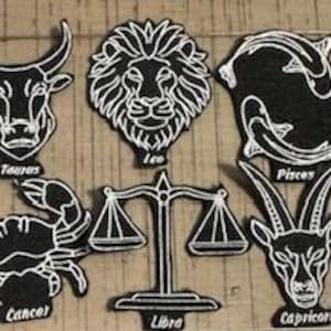 Op de afbeelding: Een verzameling zwart-witte geborduurde patches met sterrenbeeldtekens. Elke patch toont een uniek symbool en naam, waaronder Waterman, Maagd, Stier, Leeuw, Schorpioen, Kreeft, Weegschaal, Steenbok, Tweelingen, Vissen en Boogschutter.