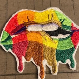 Pode incluir: Patch bordado de um par de lábios em cores do arco-íris com tinta pingando. Os lábios são delineados em preto e têm um fundo branco.