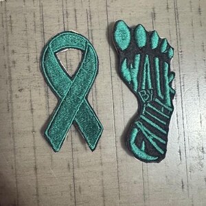 May include: Two embroidered patches: a teal awareness ribbon and a teal and black foot. The foot patch has the letters "BY" embroidered on it. Both patches have a white backing and are on a light wood-grain surface.