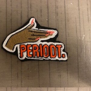 May include: A black and white embroidered patch with a gold hand with pink fingernails pointing to the right. The text "PERIODT." is in orange letters.