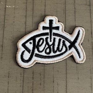 May include: Embroidered patch featuring the word "Jesus" in a stylized font within a fish shape, with a cross above. The patch has a black outline and text, with a beige border and background.