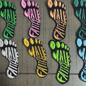Peut inclure: Écussons brodés en forme de pied, chacun avec les mots "WALK BY FAITH" dans une couleur contrastante. Les écussons sont de différentes couleurs, dont vert, rose, orange, sarcelle, blanc, jaune, vert citron et bleu. Les écussons sont sur une surface en bois.