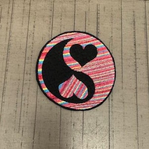 May include: A round embroidered patch with a yin yang symbol. The symbol is made up of two interlocking hearts, one black and one white. The background of the patch is a colorful striped pattern in shades of pink, red, blue, and yellow.