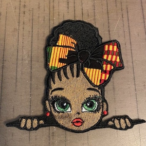 May include: Embroidered patch of a girl with black hair and a colorful bow peeking over a surface. The girl has brown skin, green eyes, and red earrings.