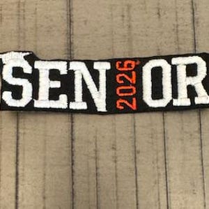 以下が含まれることがあります： 「SENIOR」の文字とオレンジ色の「2026」の年号が入った、白と黒の刺繍パッチ。卒業帽のデザインで、長方形です。
