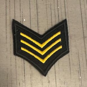 May include: Black and gold chevron patch with three gold stripes. The patch is in the shape of a downward-pointing V. The patch is on a gray wooden surface.