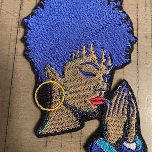 Pode incluir: Patch bordado com uma mulher de cabelo azul, olhos fechados e mãos em oração. O patch tem um brinco dourado e lábios vermelhos. O desenho é delineado em preto e tem detalhes turquesa e dourados.