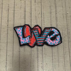 May include: A colorful embroidered patch with the word "LOVE" in a stylized font. The letters are outlined in black and filled with pink, blue, and red patterns.