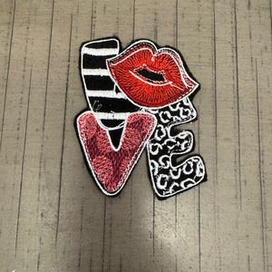 May include: A black and white embroidered patch with the word "LOVE" in a stylized font. The "O" is a red lip, the "V" is a pink heart, and the "E" is a black and white leopard print.