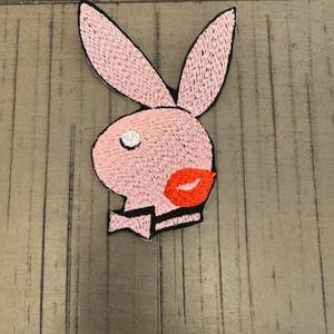 Peut inclure: Écusson brodé représentant une tête de lapin rose avec de longues oreilles, un œil blanc et des lèvres rouges. Le lapin a un col noir et blanc. Le patch est destiné aux vêtements ou accessoires.